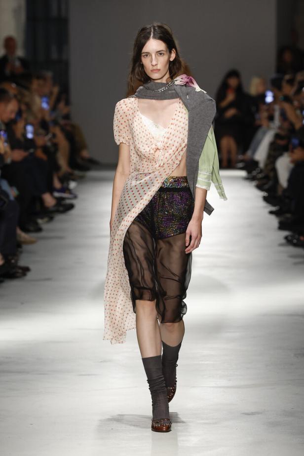 Ein Model präsentiert auf einem Laufsteg ein Outfit aus gepunktetem Kleid, glitzernder Shorts, transparentem Rock und einem über die Schulter gelegten Pullover.