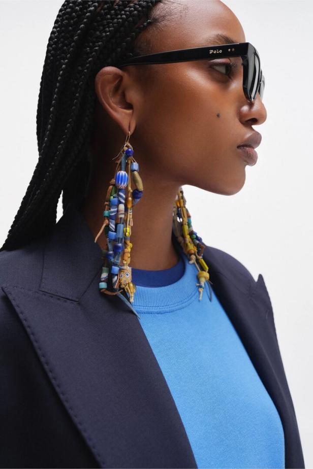 Eine Frau mit geflochtenem Haar trägt eine Polo-Sonnenbrille, ein blaues Oberteil, einen dunklen Blazer und auffällige, mehrfarbige Perlenohrringe.