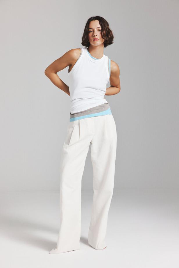 Eine Frau mit kurzem, dunklem Haar trägt ein weißes Tanktop über einem grauen und hellblauen Shirt sowie eine weite, helle Hose vor grauem Hintergrund.