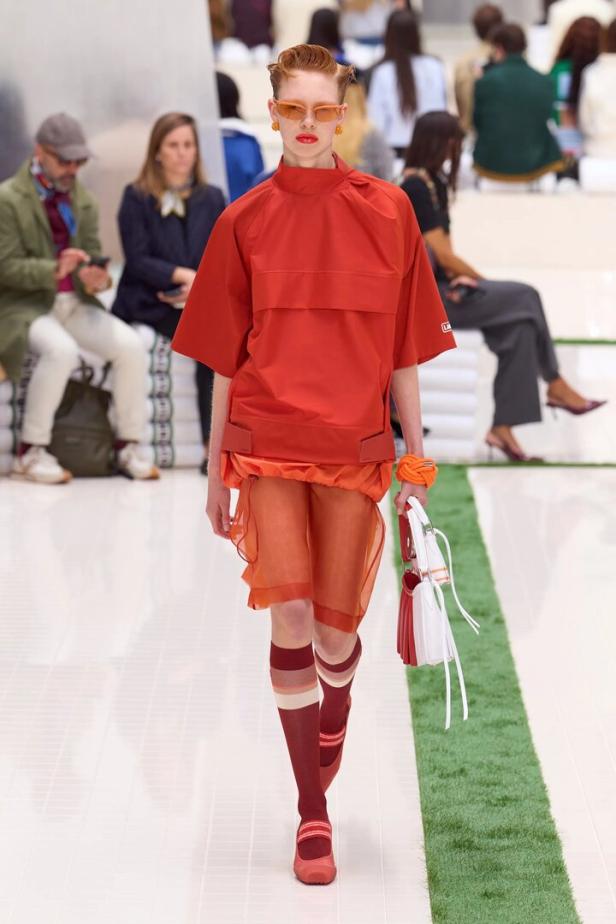 Ein Model mit orangefarbener Sonnenbrille präsentiert auf einem Laufsteg ein rotes Oberteil, einen transparenten Rock, Kniestrümpfe und eine Tasche.