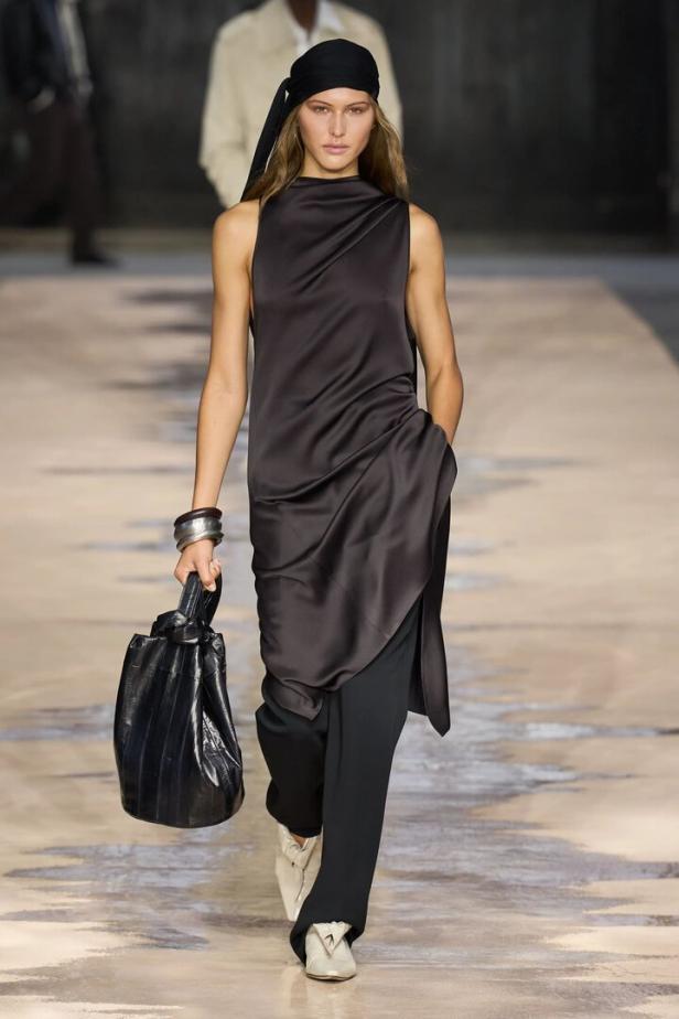 Ein Model mit schwarzem Kopftuch und ärmellosem Satinkleid läuft mit einer schwarzen Tasche über einen Laufsteg.