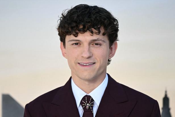 Tom Holland trägt einen dunkelroten Anzug mit einer auffälligen Spinnenbrosche am Kragen vor einem hellen Hintergrund.