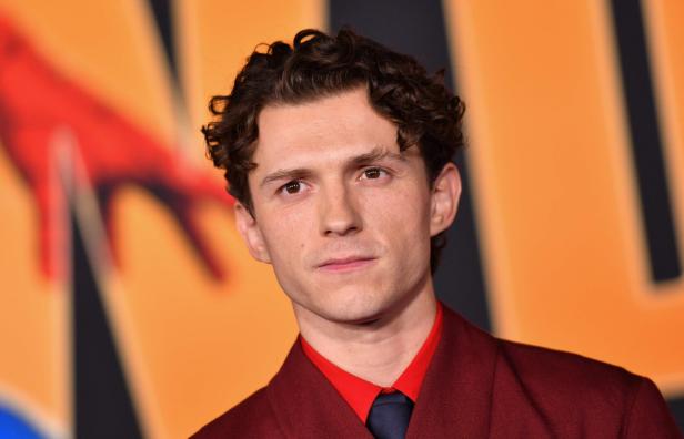 Tom Holland trägt ein dunkelrotes Sakko mit einem roten Hemd und einer dunklen Krawatte vor einem orangefarbenen Hintergrund.