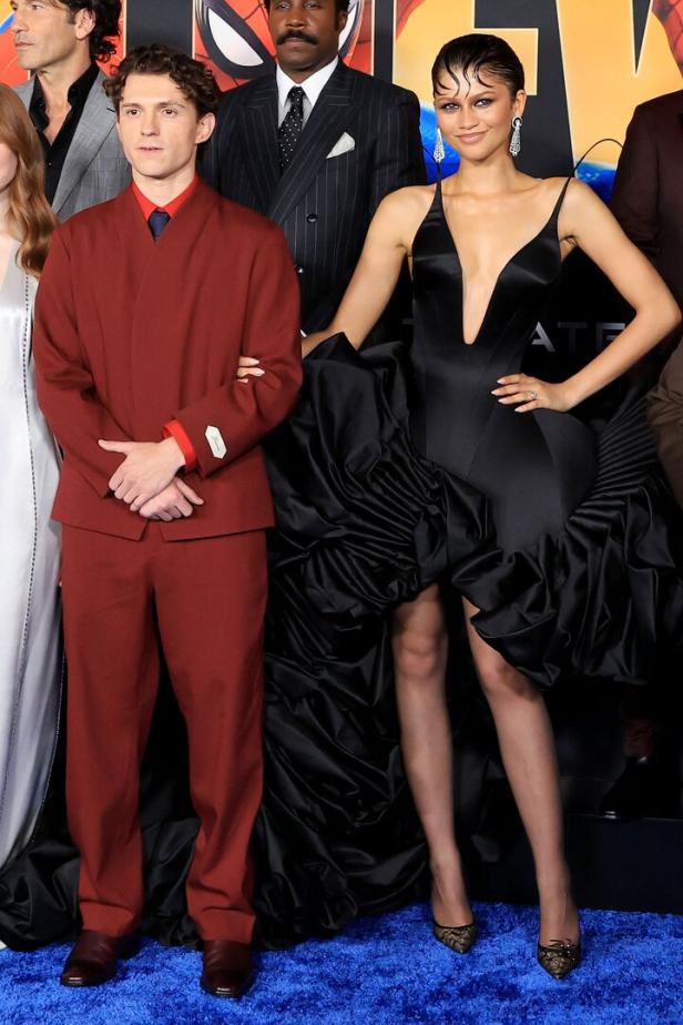 Tom Holland in einem dunkelroten Anzug und Zendaya in einem schwarzen Kleid posieren gemeinsam auf einem blauen Teppich.