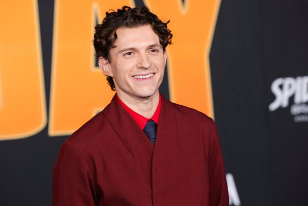 Tom Holland lächelt vor einem Hintergrund mit dem Schriftzug „Spider-Man“ in einem dunkelroten Sakko mit rotem Hemd und dunkler Krawatte.