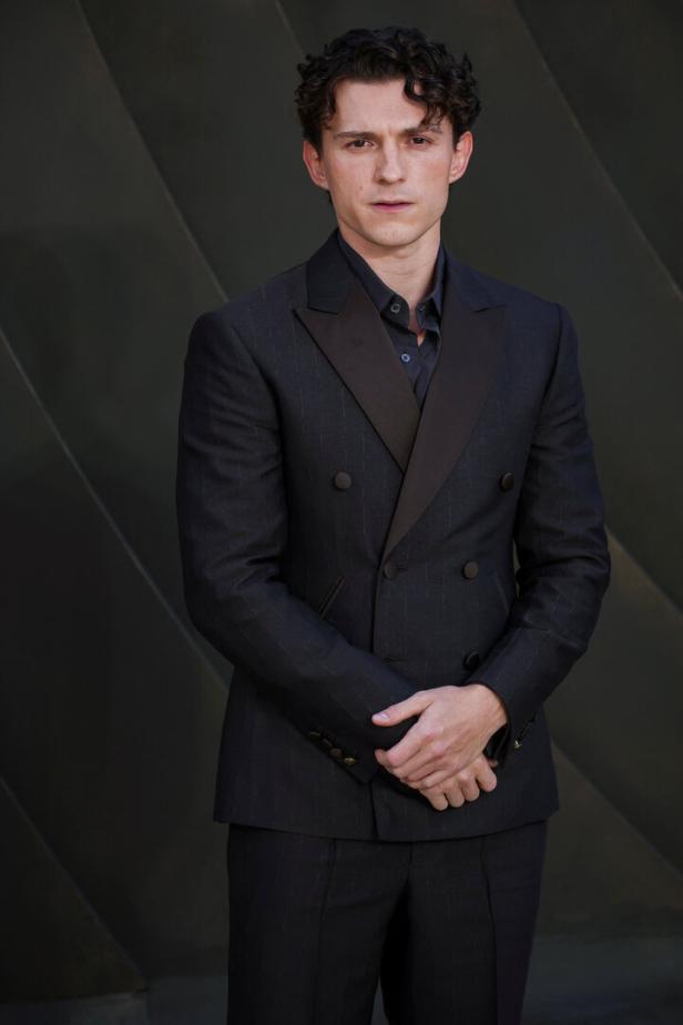 Tom Holland trägt einen eleganten, dunkelgrauen Zweireiher-Anzug vor einem schlichten, dunklen Hintergrund.