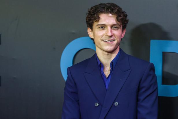 Tom Holland lächelt vor einem dunklen Hintergrund mit blauen Buchstaben in einem dunkelblauen Anzug.