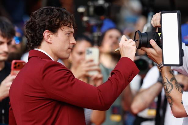 Tom Holland in einem weinroten Anzug signiert ein Kameraobjektiv vor einer Menschenmenge.
