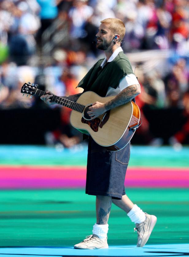 Justin Bieber spielt vor einem unscharfen Publikum auf einer Bühne mit farbenfrohem Boden eine Akustikgitarre.