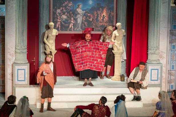 Theaterszene mit fünf verkleideten Darstellern vor rotem Vorhang und antiken Statuen, eine Figur trägt ein rotes Ziegelmuster.