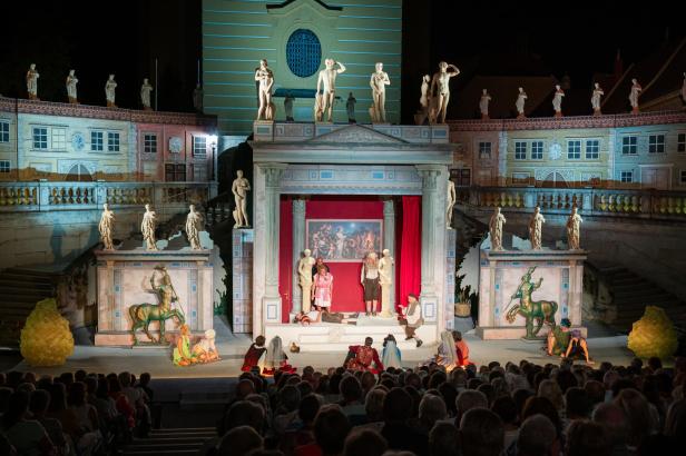 Bühnenbild einer klassischen Theateraufführung mit zahlreichen Statuen, zentralem Tempel und sitzendem Publikum im Vordergrund.
