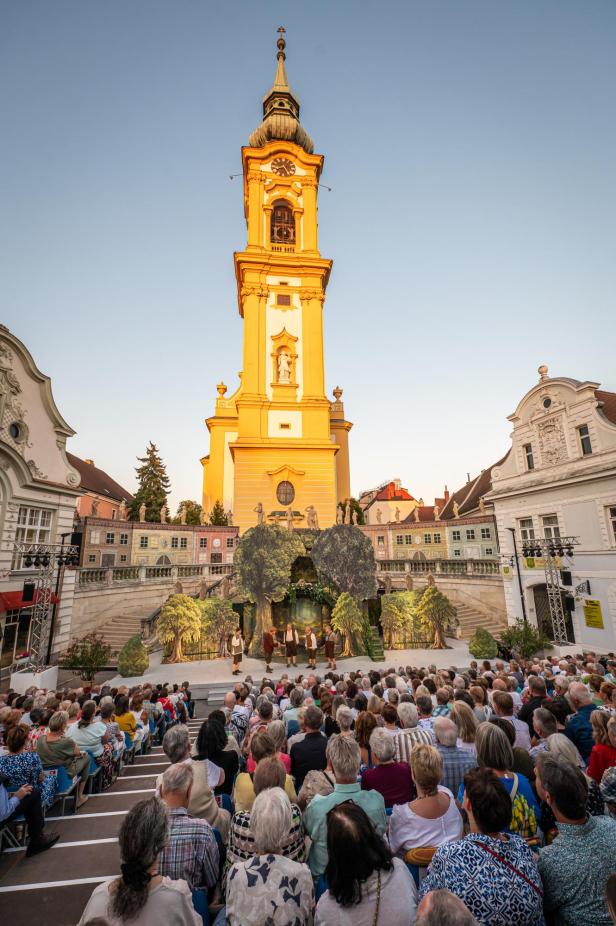 Menschen sitzen vor einer Bühne mit Theaterkulisse unter einem hohen Kirchturm bei Sonnenlicht.