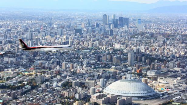 Ein Mitsubishi Regional Jet und ein Kampfjet fliegen über eine dichte Stadtlandschaft.