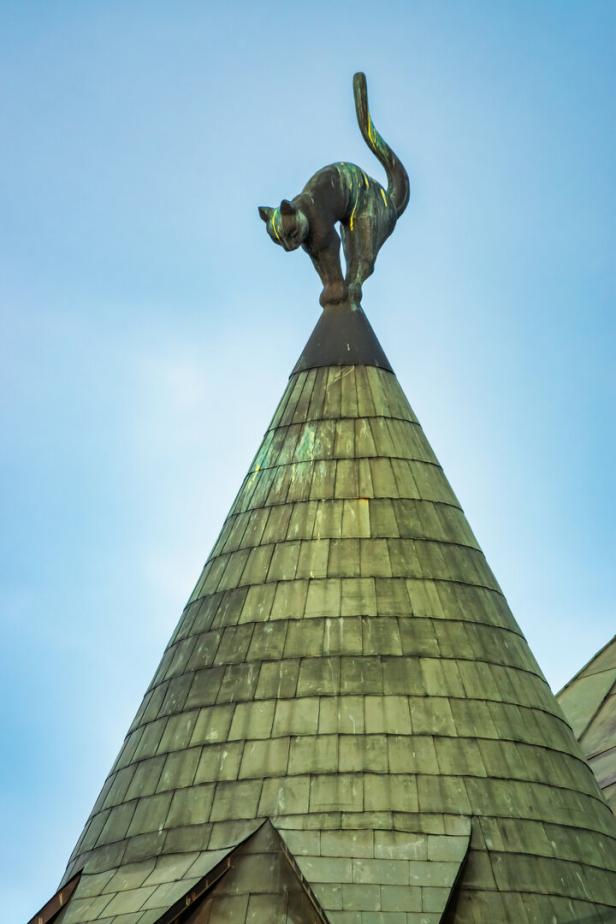 Eine bronzene Katzenfigur thront auf der Spitze eines kegelförmigen, mit Metallschindeln gedeckten Turmdachs in Riga 