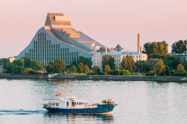 Ein Ausflugsschiff fährt auf der Düna vor der Lettischen Nationalbibliothek in Riga im warmen Licht der Abendsonne.