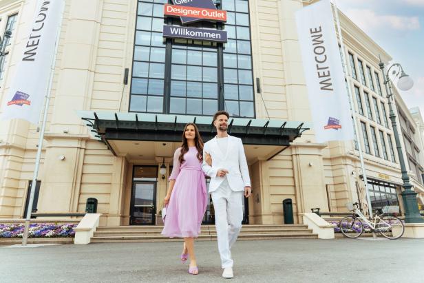 Paar vor dem Eingang des Designer Outlet mit „Willkommen“-Schildern.
