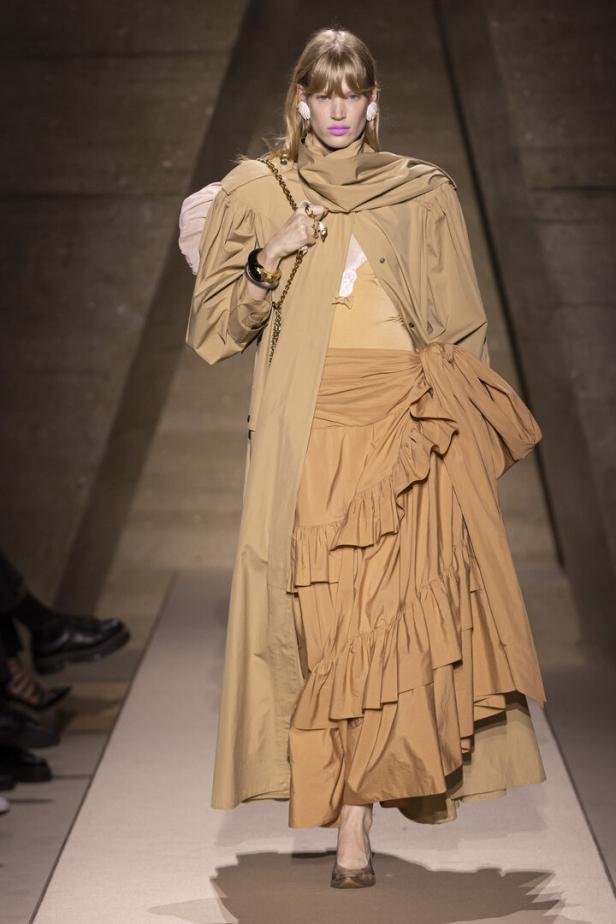 Model auf dem Laufsteg in beigefarbener, mehrlagiger Mode mit langer Jacke und Rüschrock.