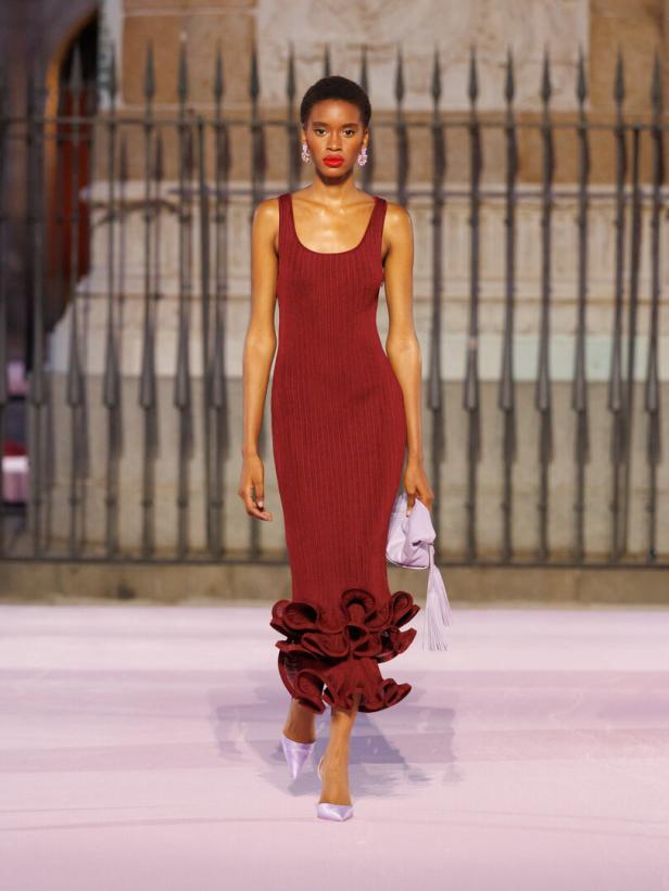 Modell läuft über den Laufsteg in einem roten, enganliegenden Kleid mit Rüschen am Saum und hält eine kleine lilafarbene Tasche.