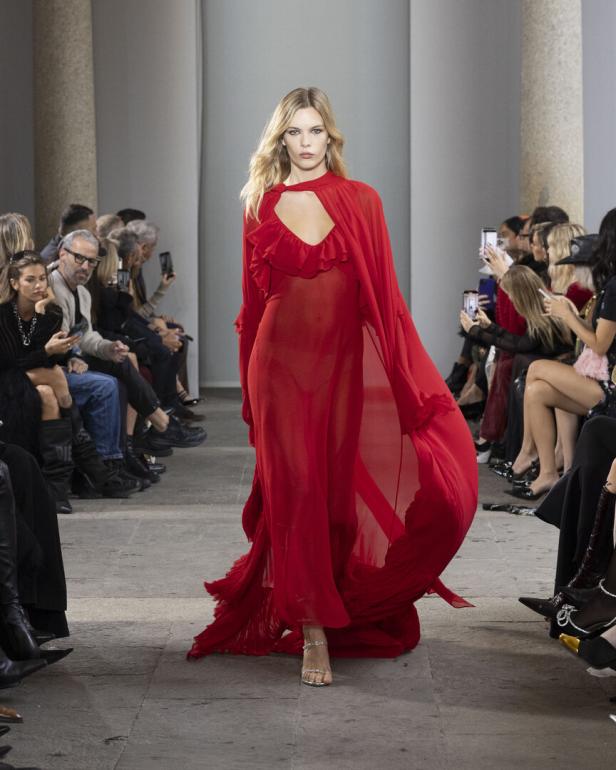 Model auf dem Laufsteg in langem, transparentem roten Kleid, flankiert von sitzendem Publikum.