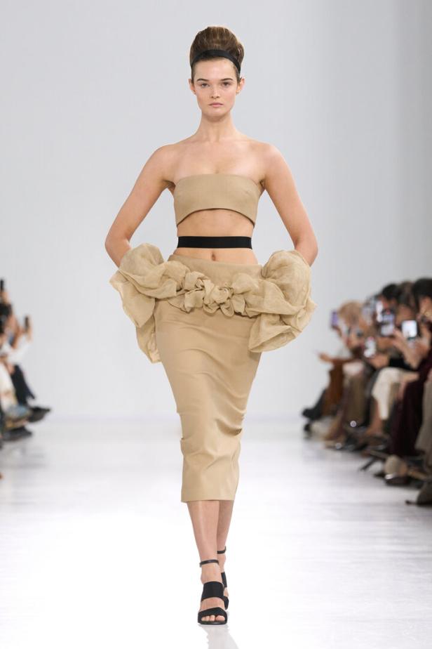 Model auf dem Laufsteg in beige-grauem Outfit mit voluminösem Rock und schwarzem Haarband.