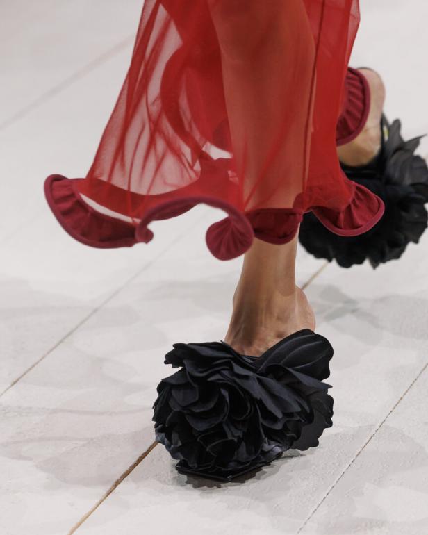Nahaufnahme von Beinen in rotem, transparentem Kleid und schwarzen Rüschen-Schuhen auf hellem Boden.