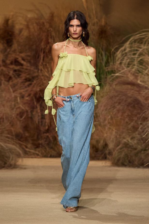 Model auf dem Laufsteg in gelbem Rüschen-Top und weiten Jeans mit Naturkulisse.