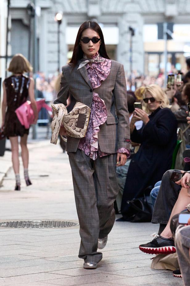 Model läuft auf einem Laufsteg in grauem Anzug mit rosa Rüschenbluse und Sonnenbrille.