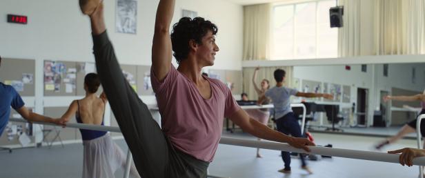 Ein junger Tänzer in einem rosa T-Shirt übt an einer Ballettstange in einem Tanzstudio, während im Hintergrund weitere Personen trainieren.