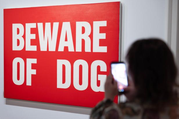Rotes „BEWARE OF DOG“-Schild in einer Ausstellung, davor eine Person mit Smartphone.