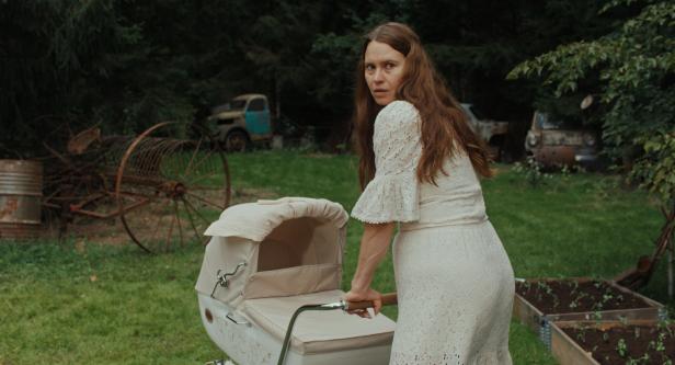 Eine Frau im weißen Kleid schiebt einen Kinderwagen im Garten, während sie sich umblickt.
