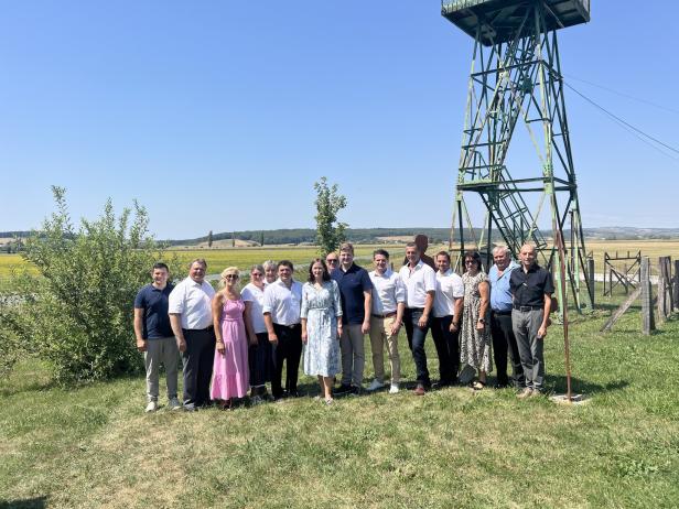 Gruppe von Menschen steht auf einer Wiese neben einem hohen Metallturm vor weiter Landschaft und blauem Himmel.