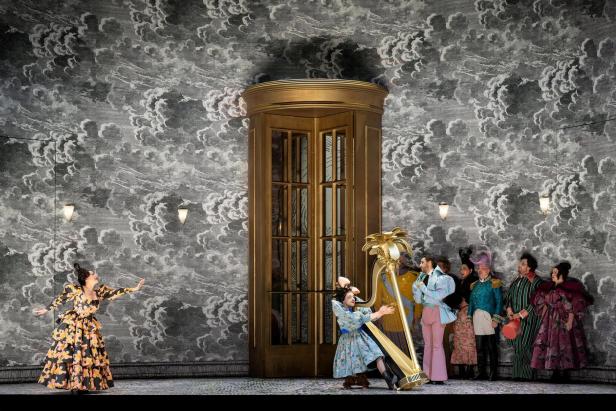 Theaterszene mit mehreren kostümierten Personen, einer Harfe und einer großen goldenen Drehtür vor wolkiger Kulisse.