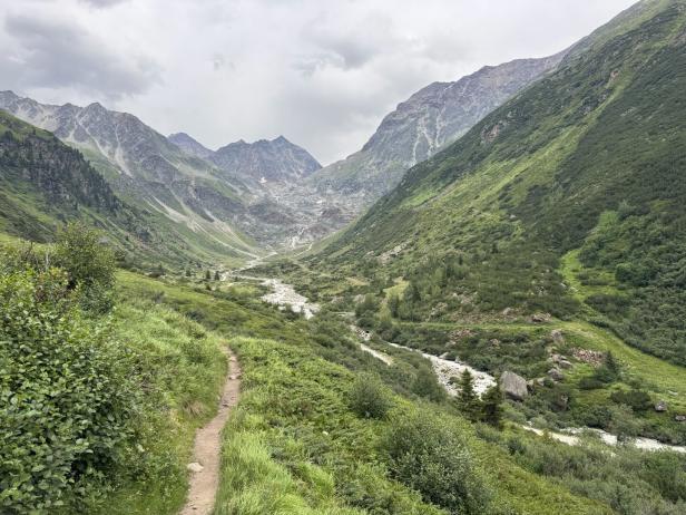 Ein schmaler Wanderweg führt durch ein grünes Gebirgstal zu einem steinigen Bachlauf vor einer imposanten Bergkulisse unter bewölktem Himmel.