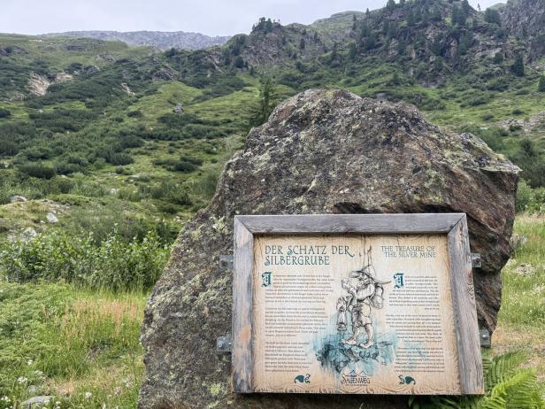 Eine Informationstafel zum Pitztaler Sagenweg über den Schatz der Silbergrube steht vor einer bergigen Landschaft.