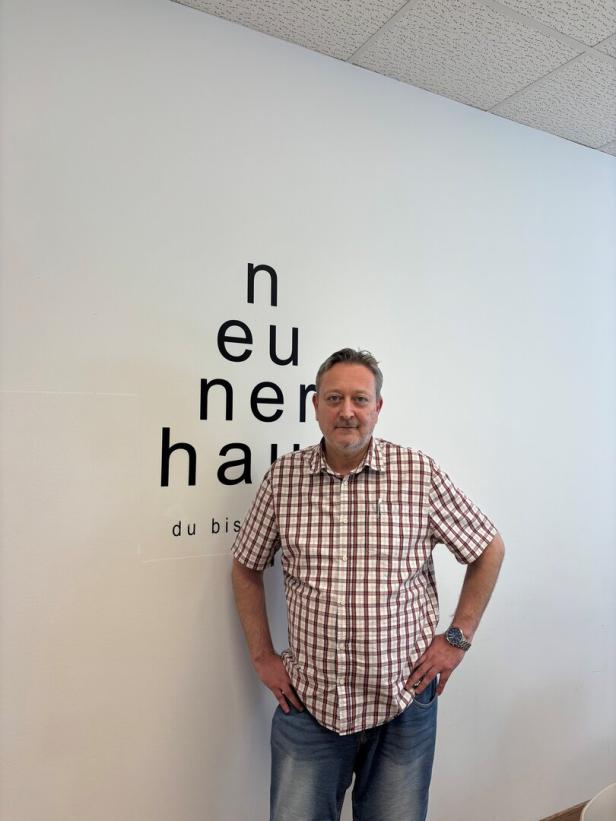 Ein Mann in einem karierten Hemd steht vor einer Wand mit dem Schriftzug „neunerhaus“.