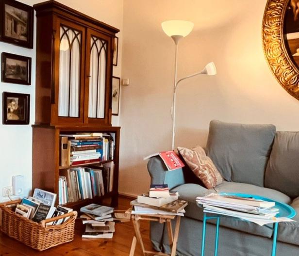 Wohnzimmer mit einem grauen Sofa, einem Bücherregal aus Holz, einer Stehlampe und zahlreichen Büchern auf Tischen und in einem Korb.