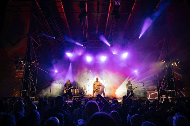 Rockband tritt auf einer Bühne mit lila Scheinwerfern und dichtem Publikum auf.
