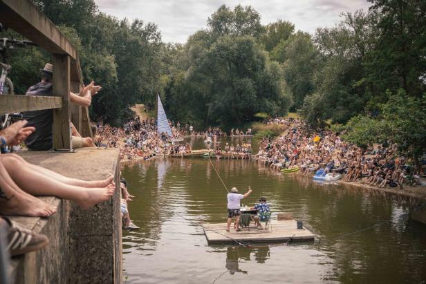 Menschen feiern dicht gedrängt an einem Flussufer, während auf einem Floß zwei Personen inmitten grüner Bäume an einem Tisch sitzen.