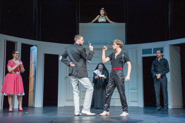 Auf einer Theaterbühne zielen zwei Männer mit Pistolen aufeinander, während eine Nonne und andere Personen zusehen.