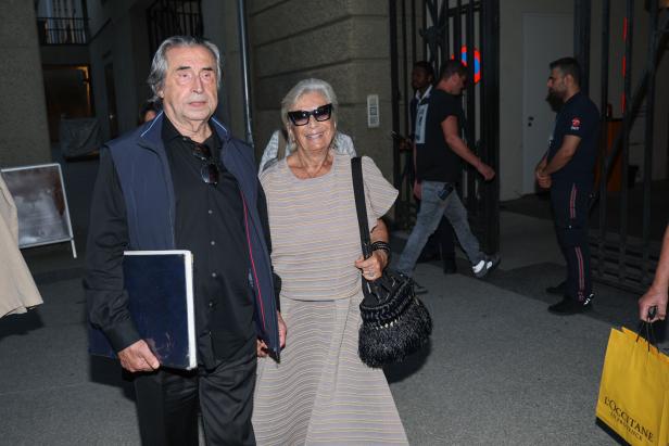Riccardo Muti und Ehefrau Cristina stehen vor einem Gebäude; er hält eine Mappe, sie trägt Sonnenbrille und Handtasche.