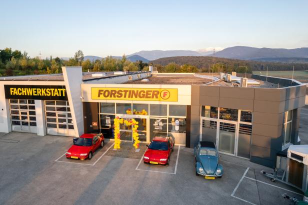 Vor einem Autohaus mit den Schildern „FACHWERKSTATT“ und „FORSTINGER“ stehen drei Autos, davor ein gelber Ballonbogen.