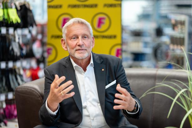 Älterer Mann im Anzug spricht gestikulierend auf einem Sofa in einem Laden, mit gelbem F‑Hintergrund.