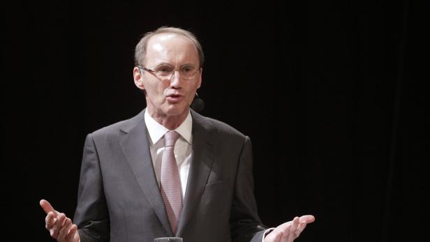 Ein Mann mit Brille und Anzug gestikuliert vor einem schwarzen Hintergrund.