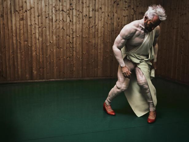 Ein muskulöser Mann in einer halb knieenden Pose trägt grünen Stoff und rote High Heels