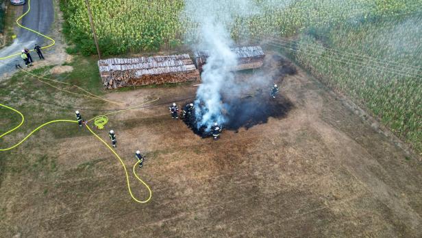 Feuerwehr löscht einen Vegetationsbrand neben Holzstapeln auf einem Feld; dichter Rauch steigt auf.