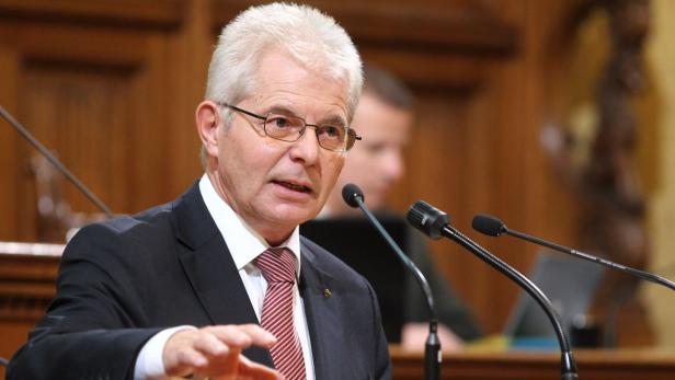 Ein Mann mit Brille spricht vor Mikrofonen in einem Saal.