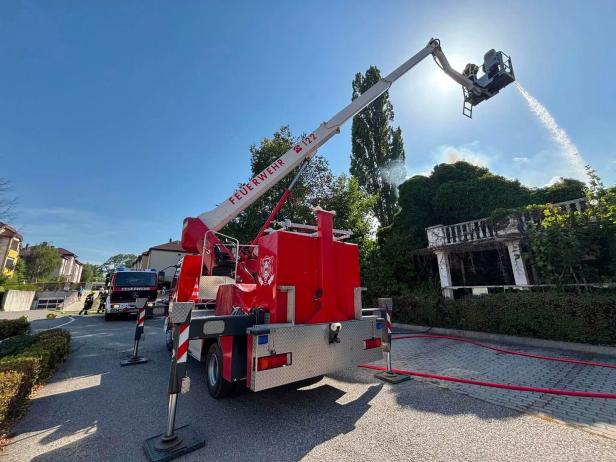 Feuerwehrmann löscht von einer Drehleiter ein brennendes Hausdach unter blauem Himmel.