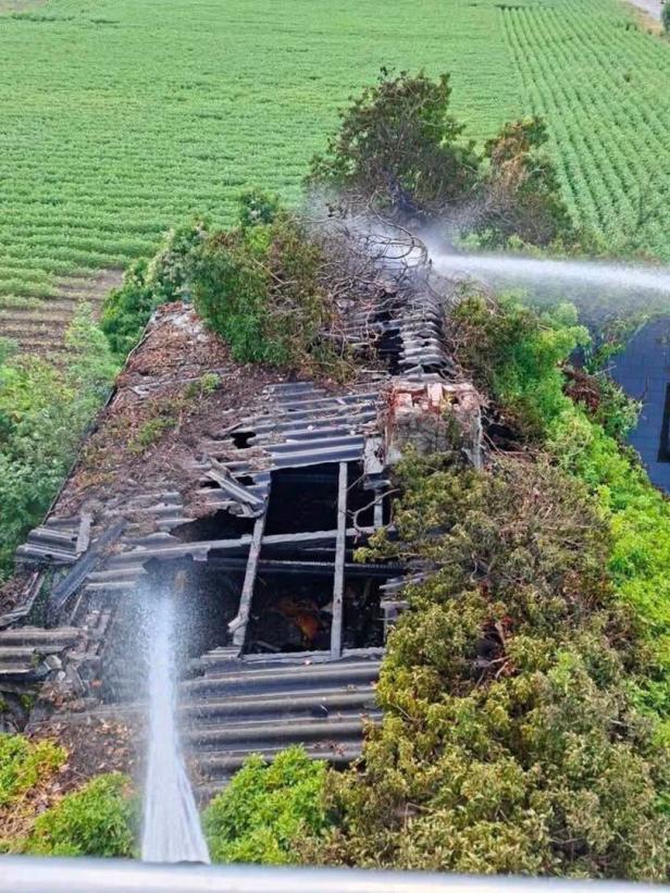 Verbranntes Dach eines Gebäudes im Grünen, über das Wasser aus zwei Strahlrohren spritzt.