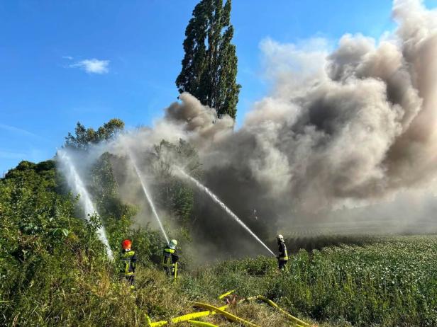 Feuerwehr bekämpft einen großen Brand auf einem Feld mit Wasserstrahlen und dichtem Rauch.