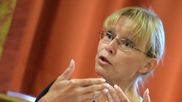 Eine Frau mit Brille gestikuliert vor einem orangefarbenen Hintergrund.